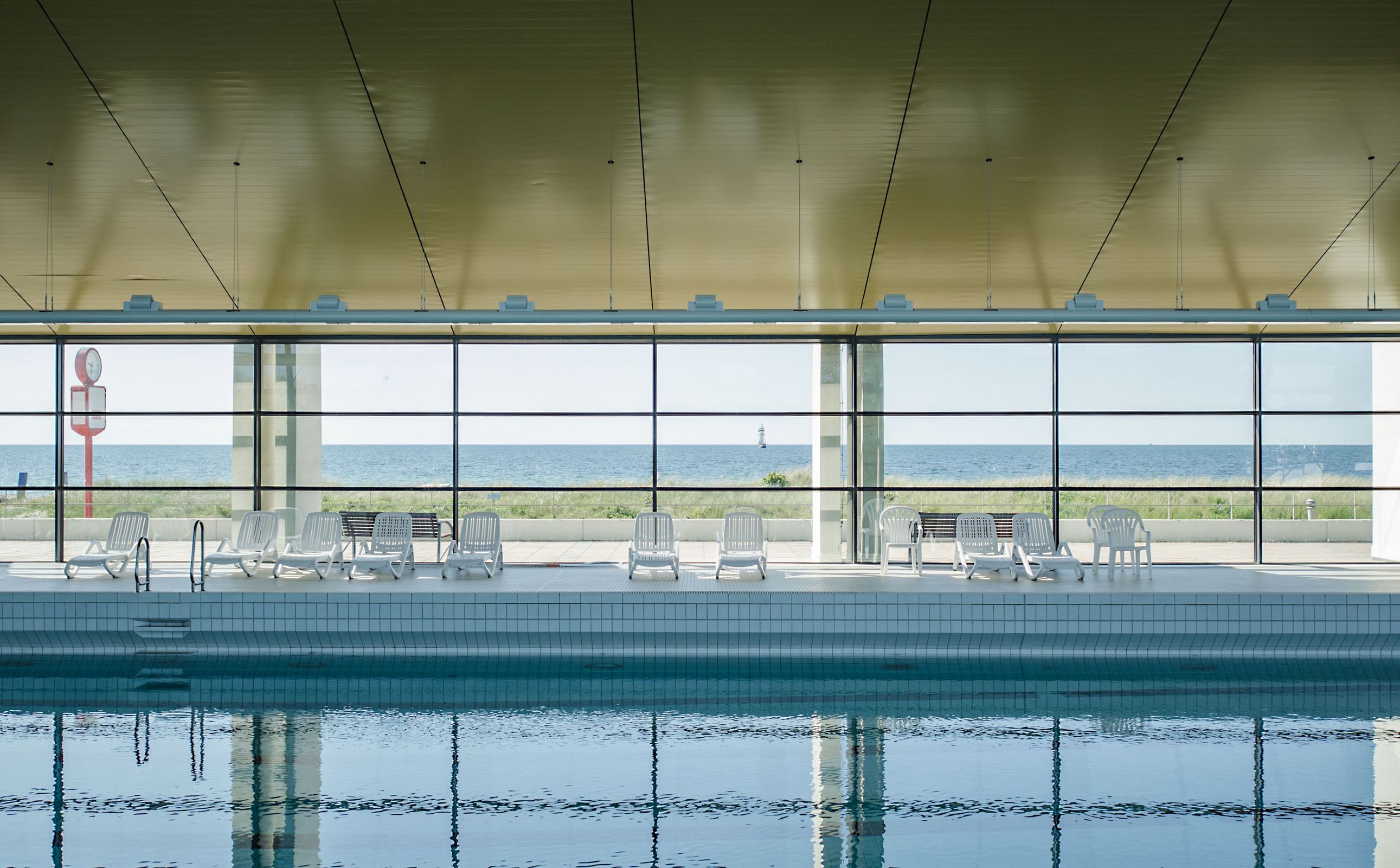 Innenansicht eines Hallenbads mit Blick durch große Fenster auf das Meer und einen Leuchtturm im Hintergrund