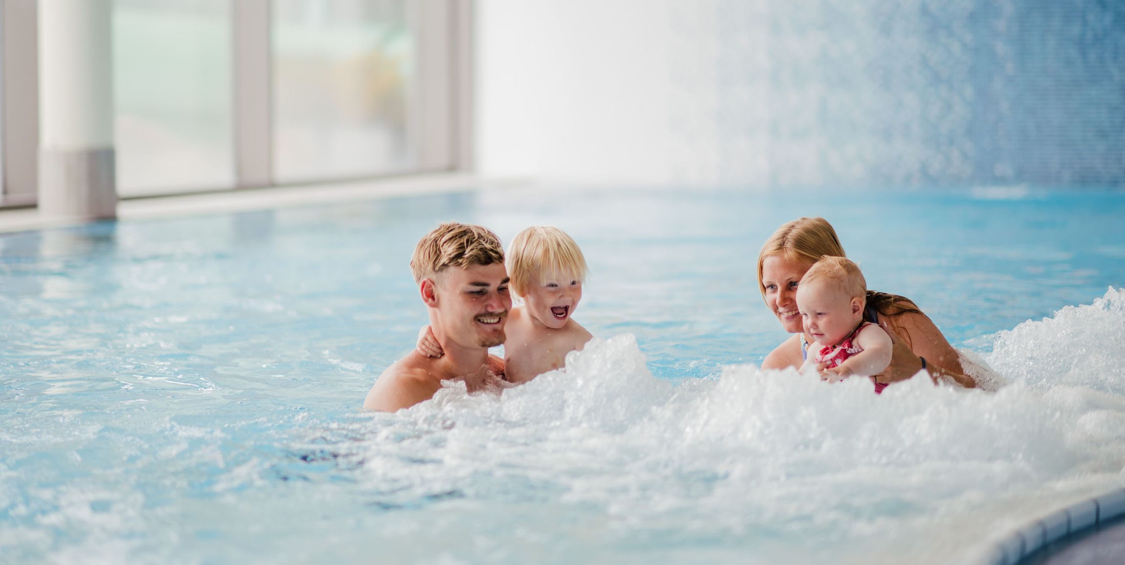 Drei Kinder spielen im Innenpool mit sprudelndem Wasser und Wasserfall im Hintergrund