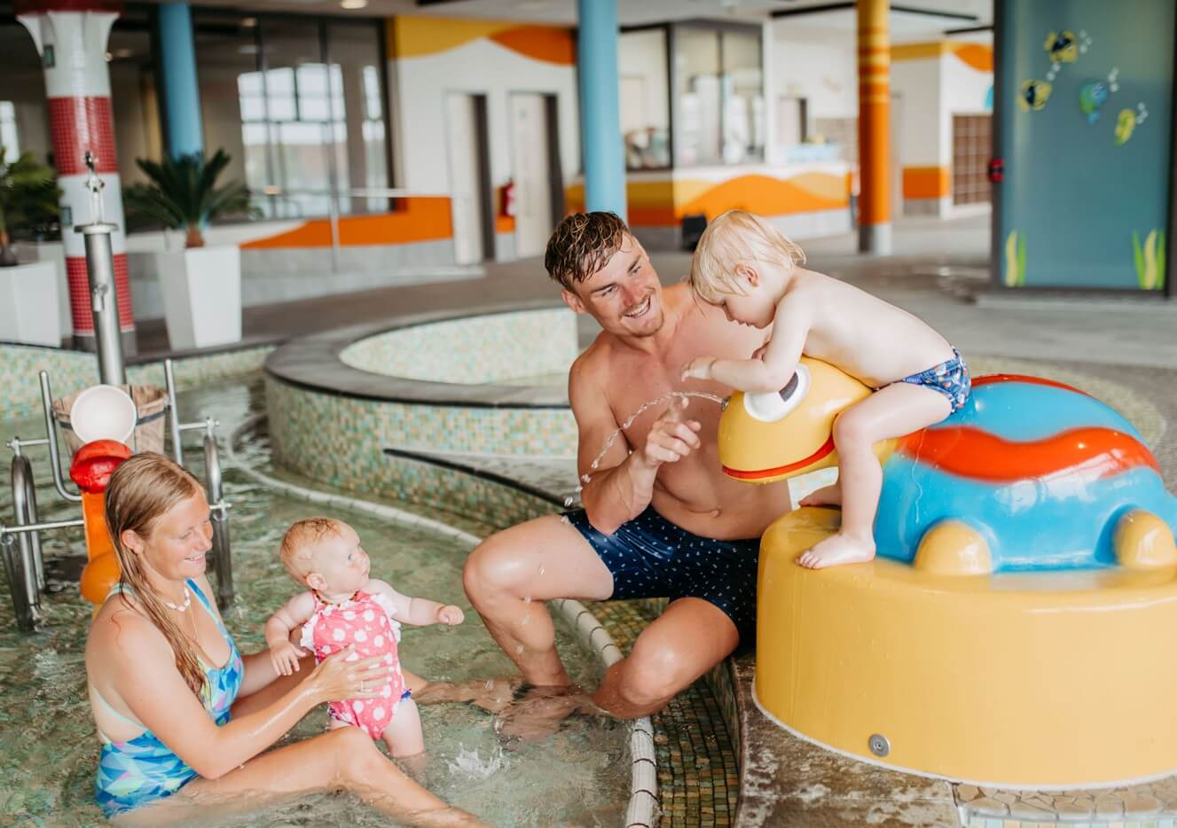 Zwei Erwachsene und zwei Kinder spielen in einem Hallenbad, ein Kind sitzt auf einer Schildkröten-Wasserrutsche.